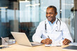 © Liubomir - Doctor in lab coat and stethoscope smiling at desk, writing on a clipboard with laptop and tablet nearby, documenting patient care in a modern clinic workspace