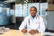 © Liubomir - Professional male physician in lab coat and stethoscope sits at a desk in a modern office, writing on a clipboard and looking confidently at the camera, symbolizing trust and medical expertise