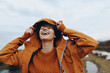 © SHOTPRIME STUDIO - Happy woman lifestyle outdoors wearing orange jacket and sweater adjusts hood in cold weather. Natural background with cloudy sky and blurred landscape. Concept of autumn, joy, and casual fashion.