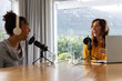 © wavebreak3 - Diverse female co-hosts talking and laughing at home studio table with mics, headphones, laptop