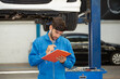 © AU USAnakul - Caucasian auto mechanic in blue uniform writing vehicle inspection checklist on clipboard while standing beneath car raised on hydraulic lift inside professional automotive repair garage workshop.