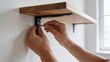 © Mahfud Picture - Detailed close-up of hands expertly securing a new wooden shelf to a pristine white wall using a screw and metal bracket, highlighting a practical home improvement task