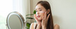 © Pormezz - Facial beauty skin health care, smile of cute asian young woman looking at mirror, hand applying moisturizer lotion on her face, female putting cream treatment before makeup cosmetic routine at home.