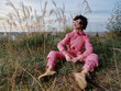 © SHOTPRIME STUDIO - Outdoor person wearing pink fashion sits among tall grasses in a field, relaxed pose with boots, wide sky, natural setting, calm mood, candid lifestyle scene for travel or editorial use