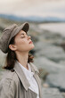 © SHOTPRIME STUDIO - Young woman with closed eyes wearing a tweed cap and coat enjoys fresh air near rocky shore. Calm serene mood in soft natural light. Concept for relaxation, nature, and peaceful moment.