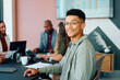 © StratfordProductions - Young businessman sitting confidently and smiling at camera while team works together in boardroom at office