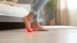 © Zabi - A person experiences sharp pain in their foot, specifically the ball of their sole, highlighted with a red glow on a light wooden floor in a bedroom setting.