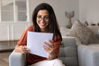 © fizkes - Beautiful Brazilian woman sit in armchair at home, smiling while reading letter notification with good news, got job offer position she wanted, received promotion, salary raise or bank loan approval