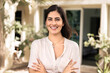 © fizkes - Happy confident young Latin homeowner woman standing outside of house, keeping hands crossed, looking at camera, smiling, posing at entrance in home patio, on backyard