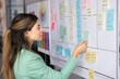 © fizkes - Busy young adult Hispanic woman Agile coach product designer adding new task to Scrum planning board creating sprint backlog thinking about process improvement structuring visual information in office