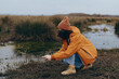 © SHOTPRIME STUDIO - Woman lifestyle concept showing a woman in an orange jacket and beanie crouching by a wetland, exploring nature with grass and water in the background during an overcast day.