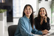 © fizkes - Happy young Asian business professional woman in glasses sitting at office table, working with positive Middle Eastern female coworker together, looking away, smiling, laughing