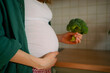 © wifesun - Pregnant woman in a home kitchen gently holding her baby bump with one hand and offering fresh broccoli with the other, symbolizing prenatal healthy eating and mindful nutrition for mom and baby