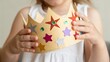 © adaptable_ Lark98A1 - A child in a white dress holds a decorative paper crown adorned with colorful star stickers.