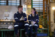 © 220 Selfmade studio - Cheerful industrial workers sitting on bench inside factory talking happily during break time relaxing with friendly conversation wearing uniform protective gear safety helmet gloves and boots
