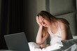© Kateryna Muzhevska - Woman with long hair sitting in bed, looking at a laptop screen with a concerned expression, illuminated by the screen light in a dimly lit room