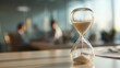 © Sutasinee - A glass hourglass standing on a desk, sand flowing. A businessman is working on a desk in a modern office background, symbol of time management, productivity, and deadline concept