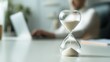 © Sutasinee - A glass hourglass standing on a desk, sand flowing. A businessman is working on a desk in a modern office background, symbol of time management, productivity, and deadline concept