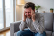 © Meow Creations - Stressed caucasian man sitting on sofa touching temples with headache pain