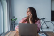 © Daniel - A happy young employee at her home workspace engages in a business call with a client, balancing digital communication and laptop work efficiently