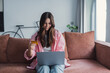 © Daniel - Happy millennial woman at home on couch, shopping from internet store using laptop and credit card, smiling as she completes purchase online, satisfied with fast and safe e-commerce experience.