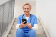 © Pixel-Shot - Happy mature female doctor with mobile phone sitting on stairs in clinic