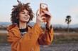 © SHOTPRIME STUDIO - A smiling woman with curly hair wearing an orange jacket takes a selfie outdoors using a smartphone. The background shows a blurred natural landscape with a tree and mountains.
