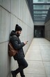 © miss irine - Man with beard wearing winter jacket, beanie, leaning against modern building wall. Holds phone, looks at screen. Brown backpack rests on shoulder. Urban city street scene in daytime, autumn vibe.