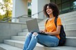 © miss irine - Young african american woman smiles widely while typing on her laptop. She sits on campus stpic with her backpack ready. Enjoying education outdoors with portable tech.