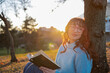 © Westend61 - Smiling woman holding book and leaning on tree trunk at sunny day