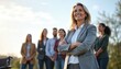 © Vadym - Successful woman leader stands with arms crossed before diverse team outdoors. One team member uses mobility aid in background. Focus on confidence and inclusion in business.