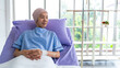 © BJ Day Stock - Asian woman cancer patient sitting alone on a hospital bed receiving saline and looking outside window in hospital ward.