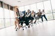 © deagreez - A group of business professionals sitting in office chairs playfully racing in a modern, bright workplace setting
