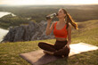 © Home-stock - Young woman in sportswear drinking water after yoga practice outside, sitting on fitness mat on hilltop at sunset, resting after outdoors workout