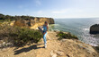 © Angelov - Woman standing confidently on a coastal cliff with a yoga mat, embracing a healthy and balanced lifestyle while enjoying outdoor wellness with the ocean and blue sky in the background