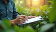 © Pete - Person in blue denim shirt writing on clipboard with pen. Surrounded by green leaves and sunlight. Focused on environmental survey or research in nature.
