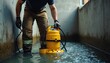 © Maryna - Worker with yellow submersible pump in flooded industrial area. Man in work pants and gloves prepares for water drainage operation. Safety gear worn for job.