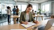 © Matt - Woman writing notes at table in collaborative office with team in background
