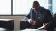 © peopleimages.com - Coffee, reading and report with business black man on sofa in lobby for management or planning. Documents, folder and information with African employee drinking beverage in workplace for review