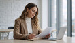 © Vyatcheslav - Professional woman reading document at office desk. Businesswoman reviewing paperwork with laptop in modern workspace. Corporate manager analyzing report