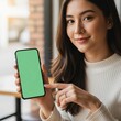 © XM7MDX - A young woman holding a smartphone with a green screen in a modern indoor setting