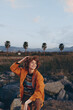 © SHOTPRIME STUDIO - Young woman smiling outdoors wearing rainbow sweater and orange jacket, sitting on rocks with natural landscape and palm trees in the background, casual lifestyle moment