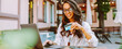 © maxbelchenko - Portrait of woman holding bank card while sitting on cozy terrace in cafe. Young woman wearing glasses and using laptop makes online purchase with debit card. Concept of online shopping.
