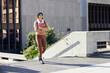 © wavebreak3 - Asian woman 30s walking across sunlit plaza wearing beanie scarf holding red notebook and backpack