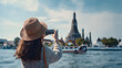 © Trendy Graphics - Woman taking a photo of a temple with a boat on the water in the background on a sunny day with a blue sky