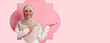 © Prostock-studio - A woman with a hijab stands in a light pink space. She points with her right hand and smiles. She wears a light sweater and appears friendly. The background is simple and decorative.