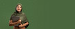 © Prostock-studio - A woman stands with a laptop in her hands. She is wearing a hijab and smiles directly at the viewer. The background is a solid green color. It appears to be daytime.
