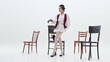 © Lustre Art Group  - Woman in poetcore outfit posing among vintage wooden chairs in white studio. Concept of fashion editorial, creative styling, boutique advertising, trend lookbook, and artistic branding visuals.