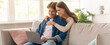 © Prostock-studio - Two people sit together on a couch in a bright living room. One person is using a laptop while the other hugs them from behind. Bright sunlight enters through the large windows.