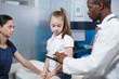 © DC Studio - Black man wearing lab coat consults with girl patient and mother in clinic office. African American physician using digital tablet while providing medical advice to caucasian woman and her daughter.